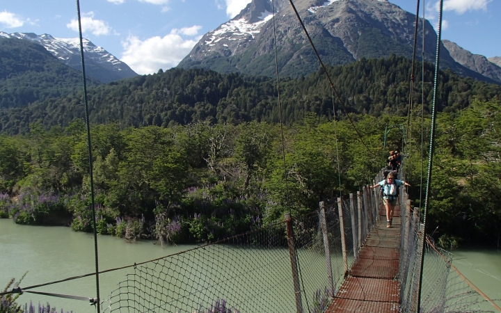 A group of people make their way over a swing bridge that hangs over a river. In the background, there are thick green trees in front of a mountain. 